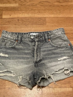 Grå jeansshorts med slitningar från Zara - Säljer ett par grå jeansshorts från Zara med slitna detaljer och råa kanter. Shortsen har fickor, knappgylf och en klassisk denimlook. Perfekta för en avslappnad och trendig stil.