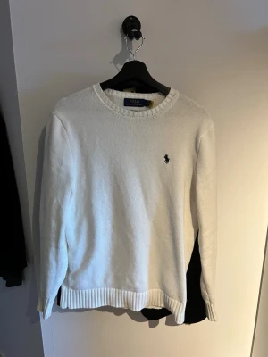 Vit stickad tröja från Polo Ralph Lauren - Klassisk vit stickad tröja från Polo Ralph Lauren med rund halsringning och svart broderad logga på bröstet. Tröjan har ribbstickade muddar vid ärmslut och nederkant. Perfekt för en clean och stilren look.