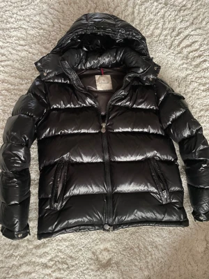 Svart dunjacka från Moncler - Snygg svart dunjacka från Moncler med glansig finish, huva och två dragkedjeförsedda fickor. Jackan har breda kanaler, hög krage och detaljerade dragkedjor med Moncler-logga. Perfekt för kalla vinterdagar och riktigt trendig look.