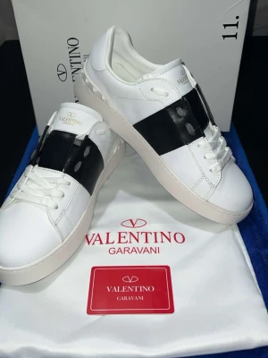 Valentino Garavani Open sneakers - Säljer ett par snygga Valentino Garavani Open sneakers i vitt skinn med bred svart läderdetalj över mitten. Skorna har rund tå, platt sula med mönstrad undersida och klassisk snörning. Perfekt för dig som gillar stilrena designer med lyxig känsla.