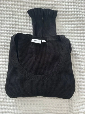 Svart ribbstickad tröja från Vila - Svart ribbstickad tröja från Vila med rund halsringning och långa ärmar. Tröjan har en smal passform och dekorativa volangdetaljer vid ärmsluten. Perfekt basplagg för lager på lager och enkel att matcha.