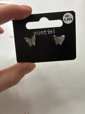 Silvriga fjärilsörhängen Montini - Snygga silvriga örhängen från Montini i form av fjärilar. Örhängena är dekorerade med små glittrande stenar som ger en lyxig känsla. Perfekta för dig som gillar söta och trendiga accessoarer. De är helt nya från urochpen