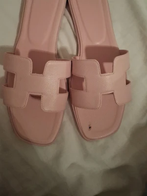 Ljusa rosa sandaler - Säljer ett par ljusrosa slip-in sandaler med fyrkantig tå och breda remmar i konstläder. Remmarna har ett stilrent, geometriskt mönster och sandalerna har en platt sula. Perfekta för sommaren och enkla att matcha med olika outfits.