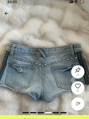 Ljusa jeansshorts med nitar och slitningar - Snygga ljusblå jeansshorts med slitna detaljer och nitar längs fickorna. Ena fickan har ett broderat mönster i blått och svart. Klassisk femficksmodell med knapp och dragkedja. Perfekta för en cool och avslappnad stil. Storlek 36 men passar mig som bär xs
