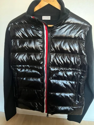 Svart pufferjacka från Moncler - Snygg svart pufferjacka från Moncler med glansig yta och stickade svarta ärmar. Dragkedjan har en röd och vit rand längs hela öppningen. Jackan har hög krage och två framfickor med dragkedja. Perfekt för kalla vinterdagar och riktigt stilren.