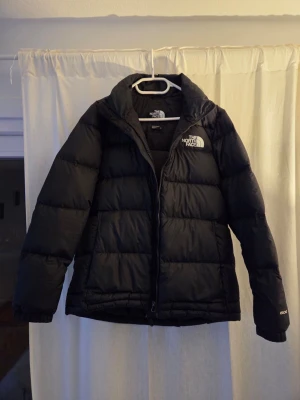 Svart pufferjacka från The North Face dam - The North Face dunjacka för dam i storlek M. Jackan är svart och har dunfyllning. Modellen har hög krage och hel dragkedja framtill. Yttermaterialet är slätt och quiltat i horisontella sektioner. Lämplig som varm ytterjacka i kallare väder. Har lite fussel inuti i jackan annars som ny.  Nypris 2800:- köpt på intersport.