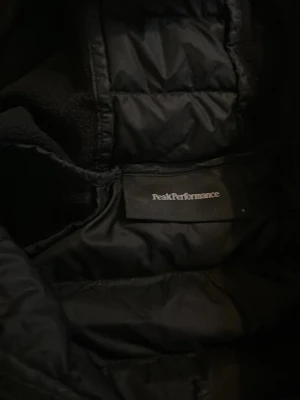 Svart pufferjacka Peak Performance - Snygg svart pufferjacka från Peak Performance med huva och quiltad design på ryggen. Jackan har släta ärmar och sidor, vilket ger en sportig look. Perfekt för kyliga dagar och enkel att matcha med olika outfits.