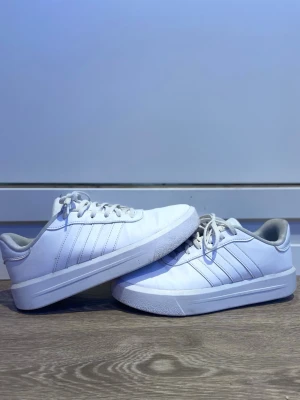 Vita Adidas sneakers med klassiska ränder - Stilrena vita sneakers från Adidas med de ikoniska tre ränderna på sidan. Skorna har lågt skaft, vit sula och snörning framtill. Tillverkade i syntetmaterial med textilfoder för extra komfort. Perfekta för en clean och sportig look.