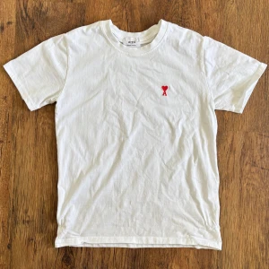 Vit t-shirt från Ami Paris - Vit t-shirt från Ami Paris med röd broderad logga på bröstet. Klassisk rund hals och korta ärmar. Tillverkad i mjuk bomull för en skön känsla. Perfekt basplagg med stilren design. SKICKAR INOM 24H✅📩