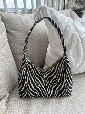 Zebramönstrad axelväska i tyg - Snygg handväska med zebramönster i svart och vitt. Väskan har en bred axelrem och stängs med magnetknapp. Insidan är beige. Perfekt för dig som vill sticka ut med ett djurmönstrat statement-plagg. Mått ca 26x15xm.