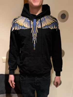 MARCELO BURLON HOODIE - Svart hoodie från Marcelo Burlon med unikt tryck av blå, gula och vita vingar över axlar och bröst. Klassisk känguruficka framtill. Perfekt statement-plagg för dig som vill sticka ut.                         (Modellen är 180cm, 72kg)