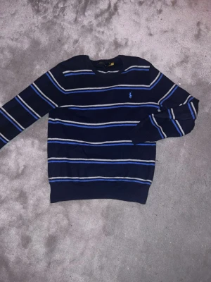 Randig stickad tröja Polo Ralph Lauren - Mörkblå stickad limiterad tröja från Polo Ralph Lauren med tunna horisontella ränder i vitt och blått. Klassisk rund halsringning och broderad logga på bröstet. Perfekt för en stilren och avslappnad look. Har ni frågor är det bara att skriva
