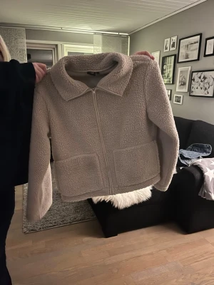 Beige teddyjacka från lager 157, stl M - Mysig beige teddyjacka från lager 157 i storlek M. Jackan har dragkedja framtill, två stora fickor och en bred krage. Perfekt för att hålla värmen och ge en chill look. Tillverkad i fluffigt teddy-material som känns supermjukt.