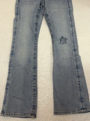 Ljusblå bootcut jeans med slitning - Säljer ett par ljusblå bootcut jeans med låg midja och slitning på ena benet. Jeansen har klassiska fem fickor och en avslappnad passform. Perfekta för en casual look och ger en cool vintagekänsla.