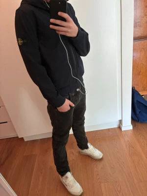 Mörkblå vindjacka från Stone Island - Snygg mörkblå vindjacka från Stone Island med huva och dragkedja framtill. Jackan har två fickor med dragkedja och den klassiska logotypen på ärmen. Perfekt för dig som vill ha en stilren och chill outfit. Perfekta vårjackan. Bra för dig som är runt 160 cm