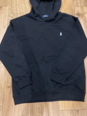 Svart hoodie från Polo Ralph Lauren - Klassisk svart hoodie från Polo Ralph Lauren med den ikoniska vita broderade loggan på bröstet. Hoodien har huva, ribbade muddar och en relaxed passform. Perfekt för dig som gillar stilrena och tidlösa plagg.