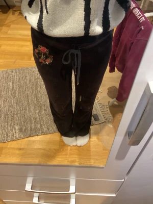 Svarta mjukisbyxor med broderi - Svarta mjukisbyxor med broderad rosdetalj på ena höften och snörning i midjan. Byxorna har raka ben och bakfickor med lock och knappar. Perfekta för en chill och avslappnad stil.