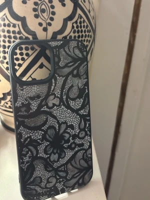 Svart mobilskal med spetsmönster - Snyggt svart mobilskal med detaljerat spetsmönster i blommig design. Skalet har en matt finish och täcker kanterna runt kameran. Passar dig som har en iPhone 15. Säljer de här fina skalet pga av att jag köpte till fel mobil.