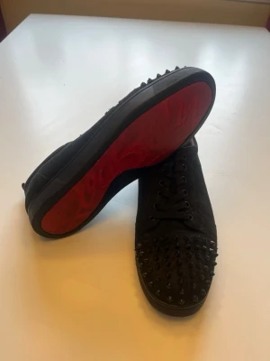 Svarta Louboutins med nitar - Säljer ett par svarta sneakers från Christian Louboutin med klassiska röda sulor och svarta nitar på tån. Skorna har snörning, låg profil och är tillverkade i mocka. Perfekta för dig som vill sticka ut med en edgy och lyxig stil.