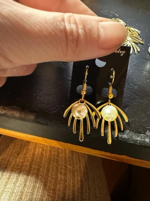 Guldörhängen med pärldetalj - Unika örhängen i guldfärgad metall med en cool, solfjäderformad design och en vit pärla i mitten. Perfekta för dig som vill sticka ut med accessoarer som känns både trendiga och eleganta. Krokarna gör dem enkla att ta på och av.