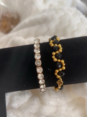 Två armband med pärlor och stenar -  två armband. Ett är guldigt med svarta och guldfärgade pärlor i ett vågigt mönster. Det andra är ett armband med klara stenar runt hela bandet som ger en glittrig look. Båda är stretch i