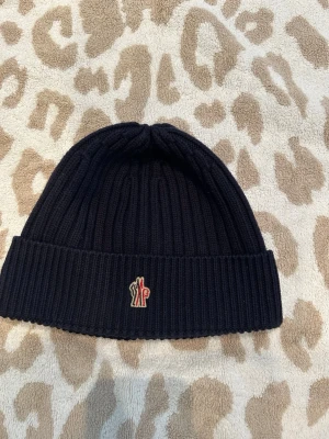 Mörkblå ribbstickad Moncler mössa - Snygg mörkblå ribbstickad mössa från Moncler med uppvikt kant och klassisk Moncler-logga framtill. Tillverkad i 100% ull vilket gör den både varm och mjuk. Perfekt accessoar för kyliga dagar och enkel att matcha med olika stilar.