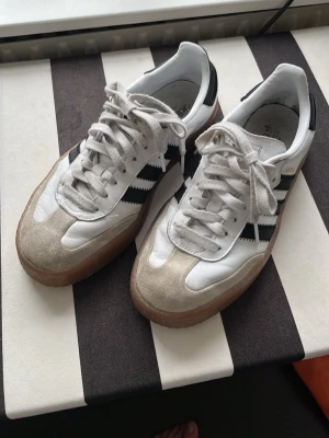 Adidas Samba vita och svarta sneakers - Adidas Samba sneakers i vitt och svart med gummisula. Skorna har tre svarta ränder på sidan, guldfärgad Samba-text och detaljer i mocka framtill. Ovandelen är i läder och de har vita skosnören. 