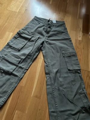 Grå cargobyxor med många fickor - Säljer ett par grå cargobyxor med bred passform och flera stora fickor på sidorna och baktill. Byxorna har klassisk cargostil med ficklock och är tillverkade i ett slitstarkt bomullstyg. Perfekta för en avslappnad streetwear-look.