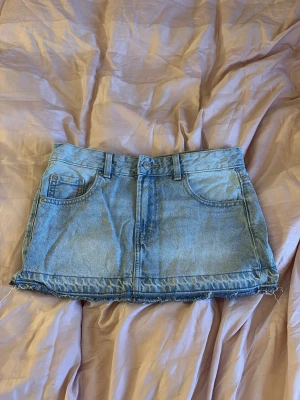 Ljusblå jeanskjol med rå kant - Trendig ljusblå jeanskjol med rå, fransig kant nertill och klassiska femficksdetaljer. Kjolen har bälteshällor och stängs med knapp och dragkedja framtill. Perfekt för en avslappnad och cool stil.