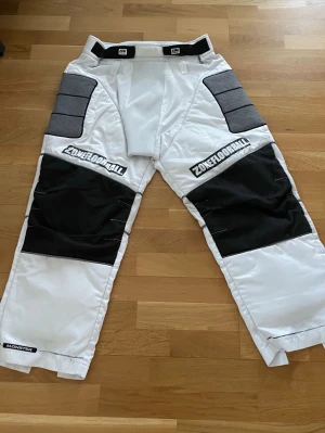 Zonefloorball vita målvaktsbyxor - Säljer ett par vita målvaktsbyxor från Zonefloorball med svarta och grå detaljer. Byxorna har vadderade partier på knän och lår, dragkedjor vid bensluten och förstärkta paneler för extra skydd. Perfekta för innebandymålvakter som vill ha både stil och funktion. 