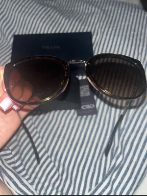 Bruna cat eye solglasögon från Prada - Snygga cat eye solglasögon från Prada med bruna bågar och mörka linser. Glasögonen har en elegant metallbrygga och kommer med originalfodral och box. Perfekta för dig som vill ha en lyxig och trendig accessoar till sommaren.