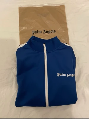 Palm Angels - Snygg blå track jacket från Palm Angels med vita ränder längs ärmarna och dragkedja framtill. Jackan har hög krage, två fickor med dragkedja och logga på bröstet. Perfekt för en sportig och trendig look.