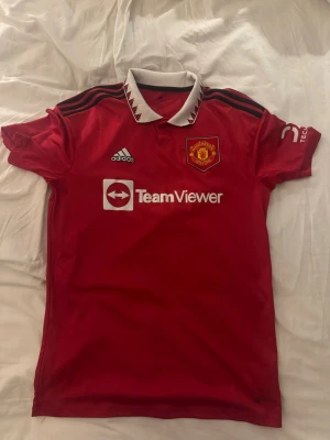 Manchester United matchtröja Adidas - Röd Manchester United matchtröja från Adidas med vit krage och svarta detaljer. Tröjan har klubbmärke på bröstet, TeamViewer som sponsortryck och klassiska Adidas-ränder på axlarna. Tillverkad i funktionsmaterial, perfekt för fotboll eller träning.