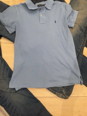 Blå pikétröja från Ralph Lauren - Snygg ljusblå pikétröja från Ralph Lauren med klassisk krage och två knappar framtill. Tröjan har den ikoniska broderade loggan på bröstet och är gjord i mjukt bomullsmaterial. Perfekt för en avslappnad men stilren look.