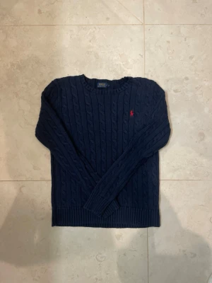 Mörkblå kabelstickad tröja Polo Ralph Lauren - Snygg mörkblå kabelstickad tröja från Polo Ralph Lauren med klassisk rund halsringning och lång ärm. Tröjan har ett rött broderat logotyp på bröstet och ribbstickade muddar vid ärmslut och nederkant. Perfekt för att hålla värmen under kyliga dagar. Står M men passar nog mer S 