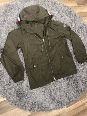Olivgrön vindjacka från Moncler - Snygg olivgrön vindjacka från Moncler med huva, storlek 1. Jackan har ett hål på insidan som du kan se på sista bilden men inget man ser när den är på.