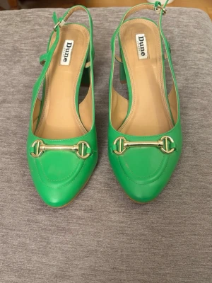 Gröna pumps med blockklack från Dune - Snygga gröna pumps från Dune med blockklack och öppen häl. Skorna har en dekorativ guldfärgad detalj framtill och justerbar rem runt hälen. Tillverkade i läder med beige innersula.