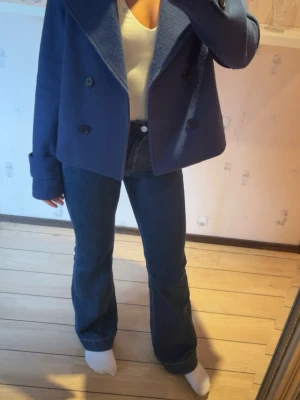 Mörkblå kappa med bred krage - Snygg mörkblå kappa med bred krage och dubbelknäppning framtill. Jackan är kortare i modellen och har stora knappar samt långa ärmar. Perfekt för dig som gillar en stilren och trendig look.