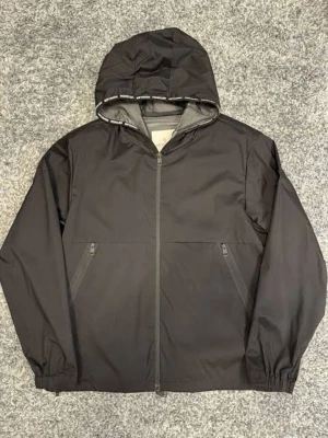 Moncler Chardon Giubbotto - Äkta Moncler Chardon Giubutto köpt i NK, Storlek 6 (moncler storlekar) , Stilren svart vindjacka med dragkedja framtill och två snedställda fickor med dragkedja. Jackan har huva med tryckt text på kanten och elastiska muddar vid ärmsluten. Perfekt för blåsiga dagar och enkel att matcha.