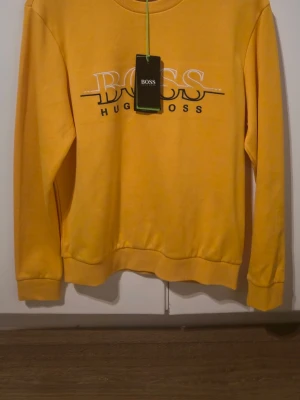 Gul sweatshirt från Hugo Boss S - Gul sweatshirt från Hugo Boss med tryckt logga i vitt och svart på bröstet. Tröjan har rund halsringning, långa ärmar och ribbade muddar vid ärmslut och nederkant. Tillverkad i mjukt material som känns skönt mot huden. Perfekt för en avslappnad och trendig stil. Aldrig använd!! Köpt på johnells! 