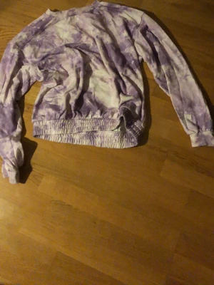 Lila tie-dye sweatshirt - En oversized sweatshirt i lila och vitt tie-dye mönster. Tröjan har långa ärmar, rund halsringning och bred resår i midjan. Perfekt för en chill och avslappnad stil. Materialet känns mjukt och bekvämt, troligen bomull eller bomullsblandning.