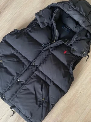 Svart dunväst från Polo Ralph Lauren - Snygg svart dunväst från Polo Ralph Lauren med avtagbar huva och dragkedja framtill. Västen har två fickor med knappar och den klassiska röda loggan broderad på bröstet. Perfekt för lager på lager och streetwear-vibe.