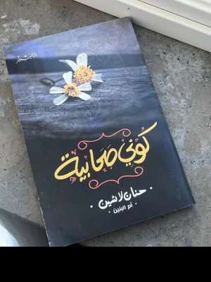 Kuni Sahiyah av Hanan Lashin - En roman med svart och lila omslag, dekorerad med två vita blommor och arabiska bokstäver i gult och vitt. Boken är skriven av Hanan Lashin och utgiven av دار تشكيل. Perfekt för dig som gillar modern arabisk skönlitteratur.