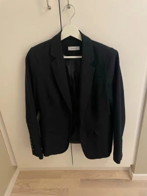 Svart kavaj från Yessica - Stilren svart kavaj från Yessica med klassisk passform. Kavajen har snygga detaljer kring kragen, och är helt oanvänd.