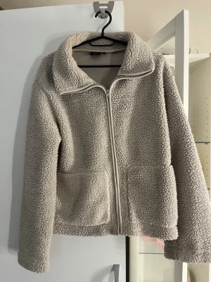 Beige teddyjacka från 157 - Mysig beige teddyjacka från 157 med dragkedja framtill och två stora fickor. Jackan har en hög krage och är tillverkad i fluffigt teddy-material som håller dig varm. Perfekt för kalla dagar och ger en avslappnad look.