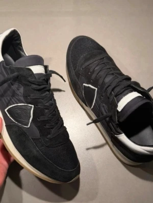  Philippe Model trainers - Stilrena svarta sneakers från Philippe Model med detaljer i vitt och grått. Ovandelen är i mocka och textil, med en ikonisk sköldlogga på sidan. Yttersulan är vit med beige inslag och skon har svarta skosnören. Perfekta för dig som gillar en clean och sportig look. Skriv vid funderingar och pris är förhandlingsbart vid snabb affär