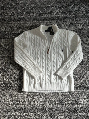 Vit kabelstickad tröja Polo Ralph Lauren - Kabelstickad vit tröja från Polo Ralph Lauren med halv dragkedja och broderad logga på bröstet. Tröjan har ribbade muddar och krage, samt lång ärm. Perfekt för dig som gillar klassisk och stilren design. Tröjan är i storlek M men passar även personer i storlek S. Hör av er om ni har några frågor eller funderingar!