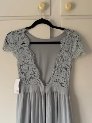 Balklänning med öppen rygg (ej använd)  - Supersöt ljusblå klänning med djup v-ringning i ryggen och spetsdetaljer upptill. Klänningen har korta spetsärmar och ett flytande kjolparti i mjukt tyg. Ej använd (prislapp kvar).