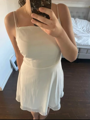 Vit kort klänning med snörning - Säljer en vit kort klänning med tunna axelband och snygg snörning i ryggen. Klänningen har en lätt utsvängd kjol och enkel, stilren design. Perfekt för sommarens alla tillfällen!