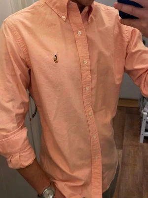 Rosa Skjorta från Ralph Lauren Oxford (Slim Fit) - Snygg Ros skjorta från Polo Ralph Lauren med klassisk button-down krage och broderad logga på bröstet. Skjortan har långa ärmar, vita knappar och är tillverkad i mjuk bomull. Perfekt för dig som vill ha en stilren och fräsch look. Storlek Small. Snabb Leverans!
